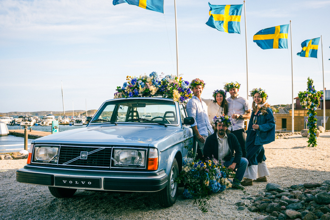 En grupp människor vid en blomsterdekorerad bil med svenska flaggor och sommarkänsla.